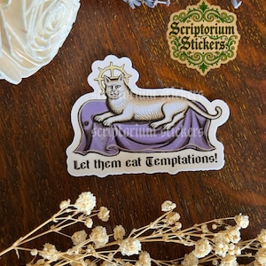 K&ouml;nnte beinhalten: Ein Aufkleber mit einer Illustration einer Katze mit Heiligenschein, die auf einem lila Kissen liegt. Der Text "Let them eat Temptations!" steht darunter. Der Hintergrund ist Holz mit floralen Elementen.