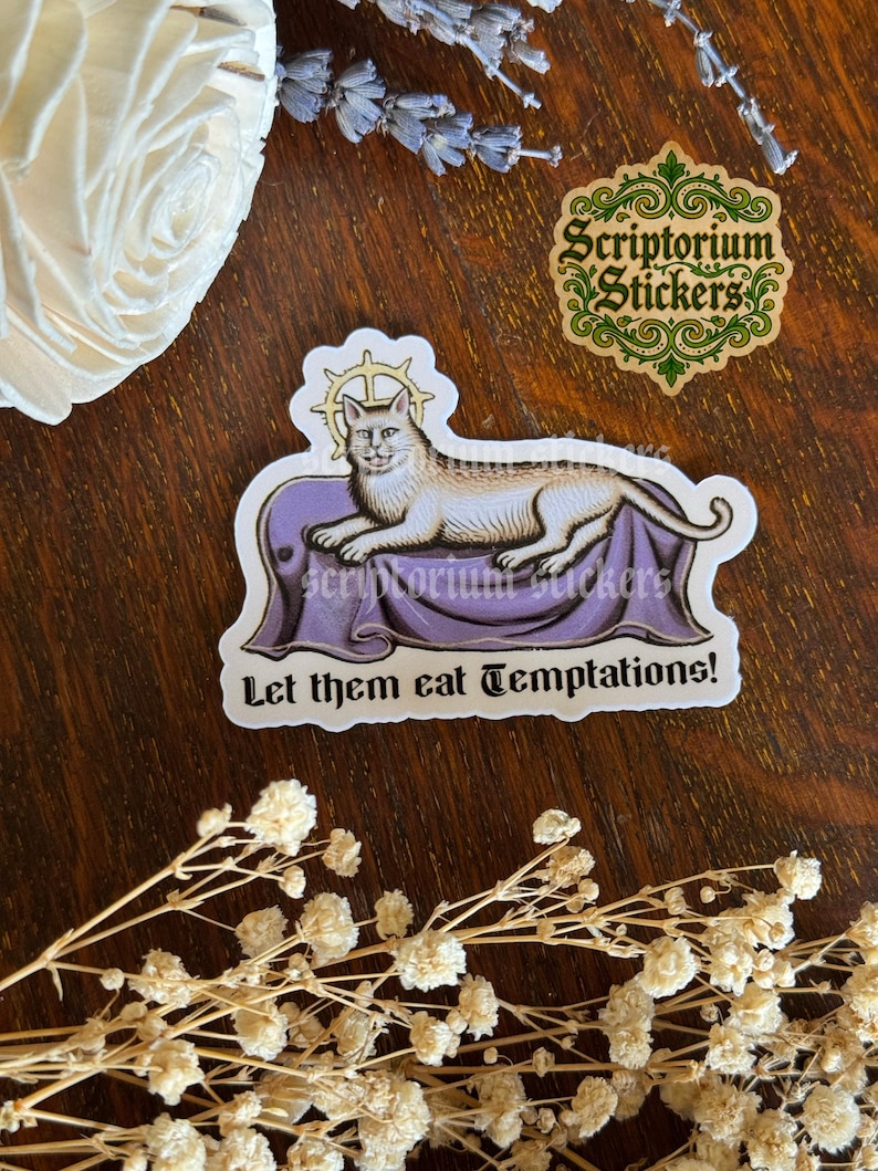 K&ouml;nnte beinhalten: Ein Aufkleber mit einer Illustration einer Katze mit Heiligenschein, die auf einem lila Kissen liegt. Der Text "Let them eat Temptations!" steht darunter. Der Hintergrund ist Holz mit floralen Elementen.