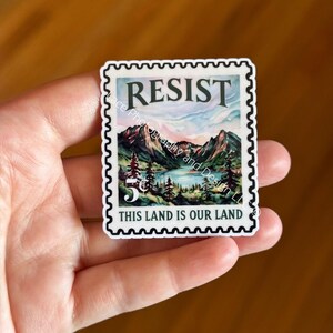 May include: A sticker in the shape of a postage stamp featuring a mountain landscape with a lake and trees. The word "RESIST" is at the top, and "THIS LAND IS OUR LAND" is at the bottom.