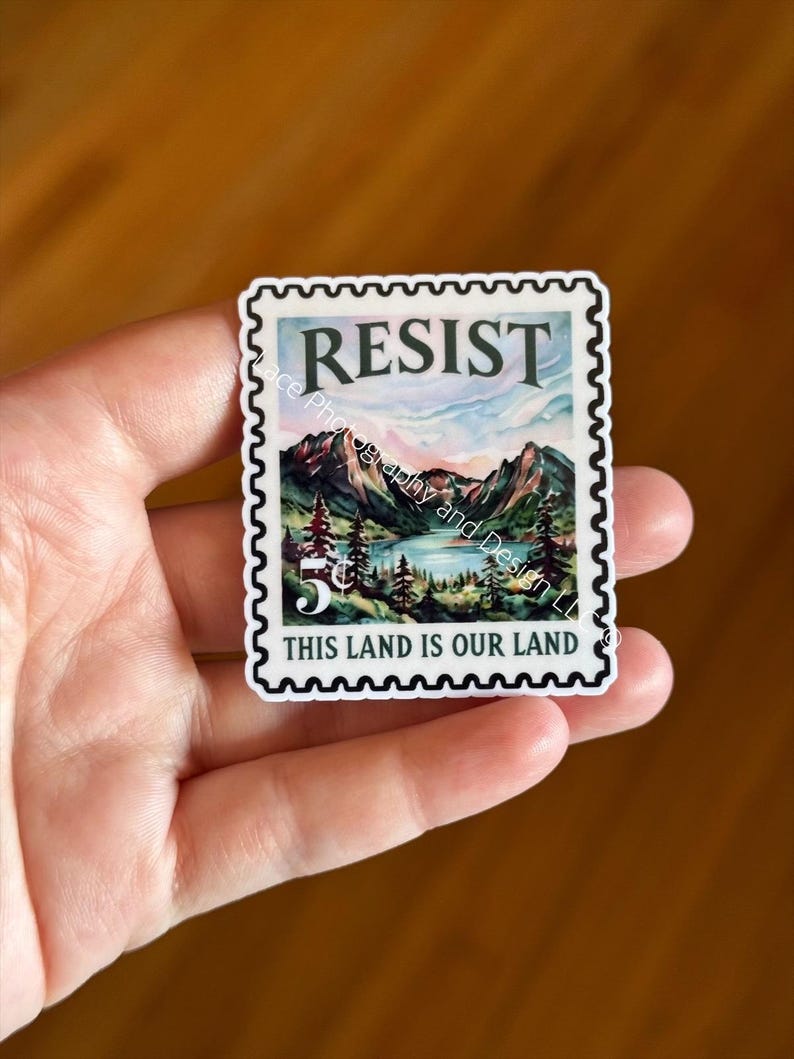 May include: A sticker in the shape of a postage stamp featuring a mountain landscape with a lake and trees. The word "RESIST" is at the top, and "THIS LAND IS OUR LAND" is at the bottom.