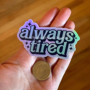 May include: A holographic sticker with the words "always tired" in a teal and black font, with the text "Lace Photography and Design LLC" below "always". A quarter is next to the sticker.