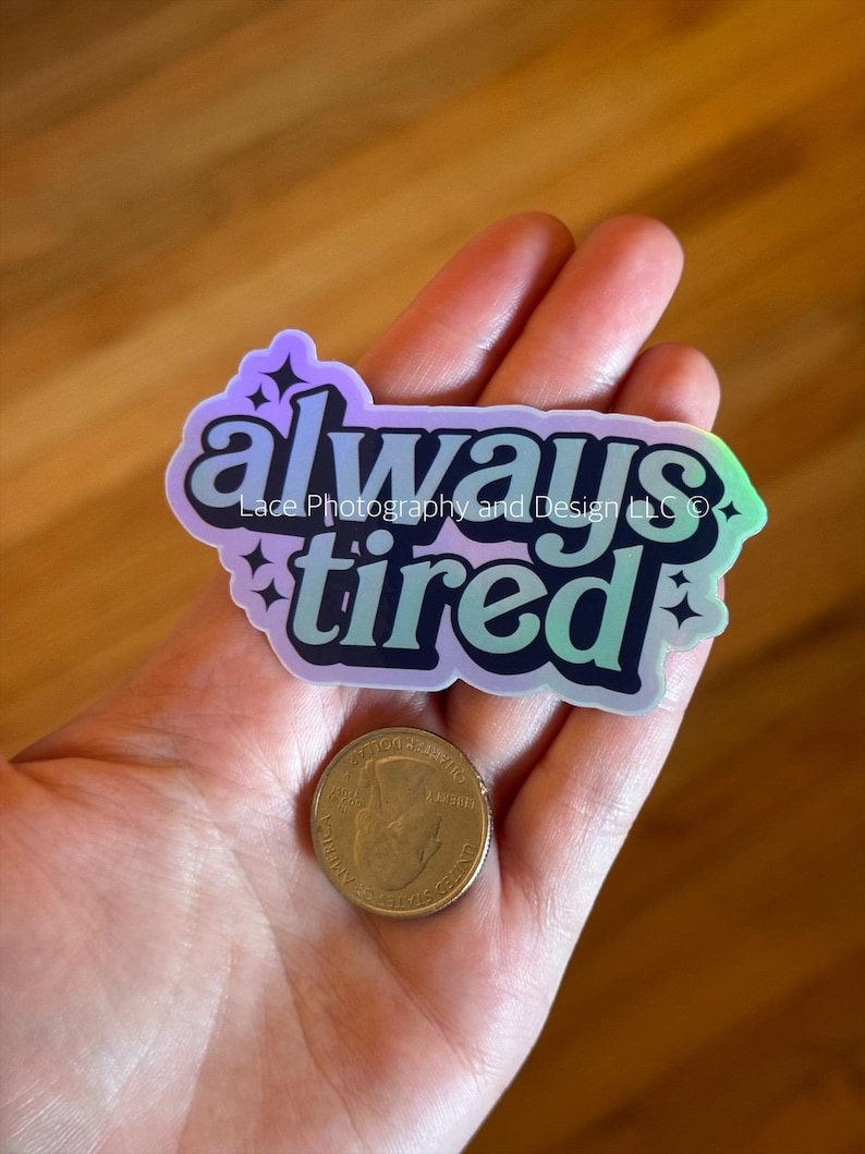 May include: A holographic sticker with the words "always tired" in a teal and black font, with the text "Lace Photography and Design LLC" below "always". A quarter is next to the sticker.