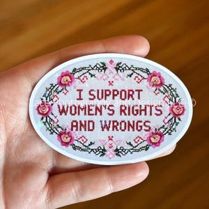 May include: Oval sticker with the text "I SUPPORT WOMEN'S RIGHTS AND WRONGS" in a cross-stitch style. The design features pink flowers and green foliage around the text, set against a white background.