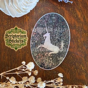 May include: An oval sticker featuring a white unicorn in a forest scene. The sticker has a dark background with floral and foliage details. A smaller sticker with the text "Scriptorium Stickers" is also visible.