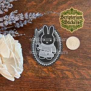 May include: A black and white sticker featuring a rabbit dressed in chainmail armor holding a sword, enclosed in an oval frame with a decorative border. A United States quarter is placed next to the sticker for scale, and the text Scriptorium Stickers is visible in the upper right corner.