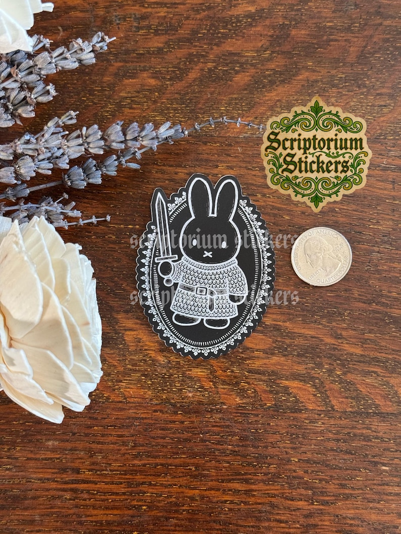 May include: A black and white sticker featuring a rabbit dressed in chainmail armor holding a sword, enclosed in an oval frame with a decorative border. A United States quarter is placed next to the sticker for scale, and the text Scriptorium Stickers is visible in the upper right corner.