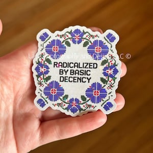 May include: A square sticker with a floral border in blue and green, featuring the text "RADICALIZED BY BASIC DECENCY" in black. The sticker is held in a hand.