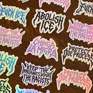 May include: A collection of stickers with bold, stylized text in various colors. Phrases include "Abolish ICE", "Protect Due Process", and "Keep the Immigrants Deport the Racists". The stickers have a glossy finish and are arranged on a wooden surface.