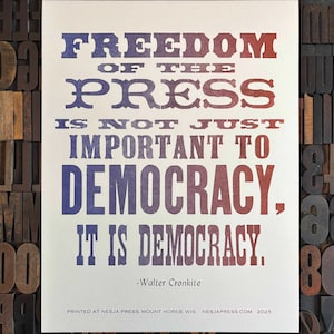 May include: A print with the quote "FREEDOM OF THE PRESS IS NOT JUST IMPORTANT TO DEMOCRACY. IT IS DEMOCRACY." in blue and red text. The print is on a cream-colored background. The quote is attributed to Walter Cronkite.