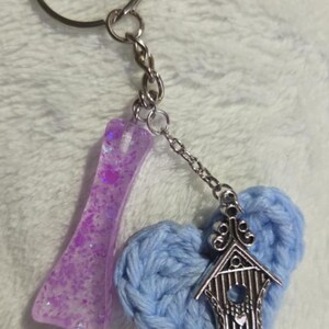 May include: A handmade keychain featuring a purple resin charm with glitter, a light blue crocheted heart, and a silver-colored birdhouse charm. The keychain is attached to a silver-colored key ring.