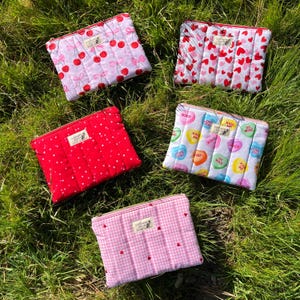 以下が含まれることがあります： 緑の草の上に、さまざまな模様のキルティングコスメティックバッグが5つ並んでいます。模様には、さくらんぼ、ハート、そして「Kiss Me」や「Love You」などのフレーズが書かれた会話用のハートが含まれています。各バッグにはジッパー開閉と小さなラベルが付いています。