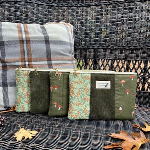 Könnte beinhalten: Drei olivgrüne Reißverschlusstaschen mit goldenen Reißverschlüssen. Eine Tasche hat ein Blumenmuster, eine andere eine Cordstruktur und die dritte ein Pilzdesign. Im Hintergrund befindet sich ein Kissen mit Karomuster.