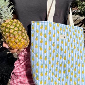Puede incluir: Un bolso tote acolchado con un estampado de rayas azules y blancas con piñas amarillas. El bolso tiene una correa de color crema. Se sostiene una piña fresca junto al bolso. La persona lleva una camiseta negra y pantalones a cuadros rojos y blancos.