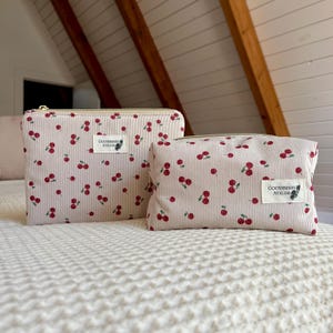 May include: Two light pink corduroy zippered pouches with a cherry pattern. The pouches have a vertical striped texture and a gold zipper. Each pouch has a white label with the text "GOOSEBERRY ATELIER" and a small cherry illustration.