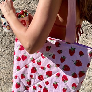Puede incluir: Un bolso tote acolchado con fondo rosa y un estampado de fresas rojas. El bolso tiene una correa rosa claro y una etiqueta que dice "Gooseberry Atelier". El bolso se sostiene al aire libre.