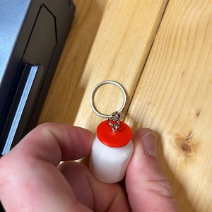 Può includere: Un portachiavi cilindrico bianco con un tappo rosso. Il portachiavi è attaccato a un portachiavi argentato. Lo sfondo è una superficie di legno.