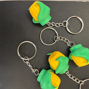 May include: Several corn-shaped keychains. Each keychain features a yellow corn cob with green husks. The corn is attached to a silver chain and a silver key ring. The keychains are on a dark surface.