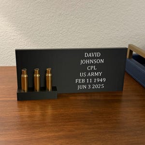 May include: A black memorial plaque with the name "DAVID JOHNSON" and military information, along with three brass bullet casings. The text includes birth and death dates. The plaque is on a wooden surface.