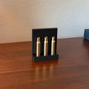 May include: A black rectangular display stand holding three gold-colored bullet-shaped objects. The stand is on a dark wooden surface, with a blue and white decorative plate and a lamp in the background.