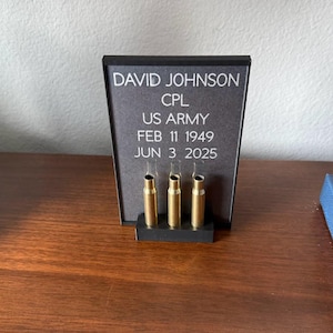 May include: A memorial display featuring a black frame with the name "DAVID JOHNSON", rank "CPL", and "US ARMY". The dates "FEB 11 1949" and "JUN 3 2025" are also displayed. Three brass bullet casings are set in a black base.