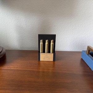 May include: A memorial display featuring three brass bullet casings in a black stand. The stand has a gold plaque with the text "DAVID JOHNSON, CPL, US ARMY, FEB 11 1969, JUN 3 2020". The display is on a wooden surface.