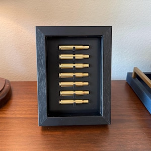 May include: A framed display featuring seven golden bullet casings arranged vertically against a black background. The casings are evenly spaced within a black wooden frame, creating a unique decorative piece.