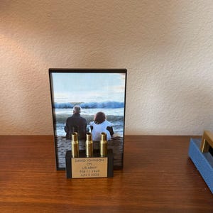 May include: A framed photo with a beach scene and three gold-colored bullet casings. The photo features two people looking at the ocean. Below the photo is a black base with text that reads "DAVID JOHNSON, US ARMY, FEB 11 1949, JUN 3 2025."