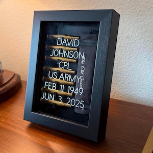 May include: Black framed shadow box displaying military memorabilia. The text reads: "DAVID JOHNSON, CPL, US ARMY, FEB 11 1949, JUN 3 2025." Includes a black fabric with the text "The Old Guard."