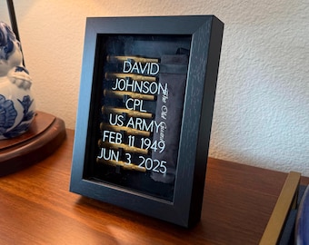 May include: Black framed shadow box displaying military memorabilia. The text reads: "DAVID JOHNSON, CPL, US ARMY, FEB 11 1949, JUN 3 2025." Includes a black fabric with the text "The Old Guard."