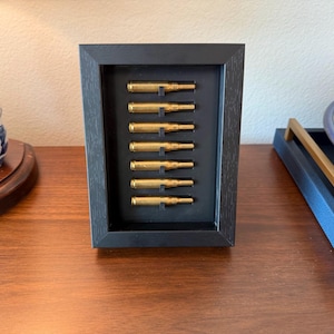 May include: A framed display featuring eight brass bullet casings arranged vertically against a black background. The frame is black and rectangular, creating a minimalist aesthetic. The casings are evenly spaced, showcasing a unique decorative item.