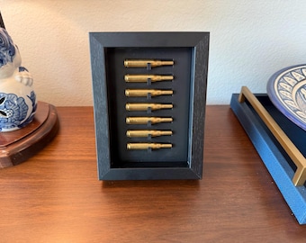May include: A framed display featuring eight brass bullet casings arranged vertically against a black background. The frame is black and rectangular, creating a minimalist aesthetic. The casings are evenly spaced, showcasing a unique decorative item.