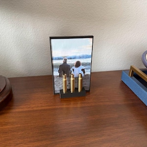 May include: A black framed photo with a picture of two people sitting on a beach. Three gold-colored bullet casings are in front of the photo. The photo frame is on a dark brown wooden surface.