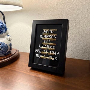 May include: A framed memorial with the name "DAVID JOHNSON", rank "CPL", and "US ARMY" displayed. The frame includes the birth date "FEB 11 1949" and the date of passing "JUN 3 2025". The frame is black and sits on a wooden surface.