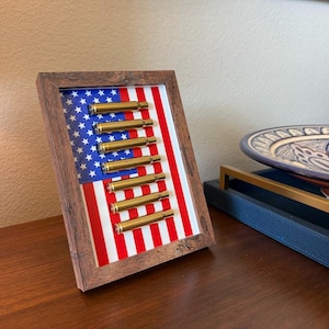 May include: A framed artwork featuring an American flag design with brass bullet casings arranged vertically. The frame is made of dark wood, and the flag has red and white stripes with a blue field of white stars.