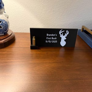 May include: A black desk sign with the text "Brandon's First Buck 11/15/2025" and a white deer silhouette. A brass bullet casing is displayed next to the sign. The sign is on a wooden surface.