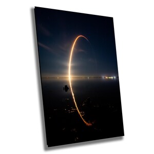 May include: A long exposure photograph of a rocket launch at night. The rocket's trail of fire is visible in the sky, creating a bright arc over a body of water. The water reflects the light from the rocket, creating a mirror image of the trail.