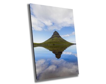 May include: A green mountain with a pointed peak reflected in a still body of water. The sky is blue with white clouds.