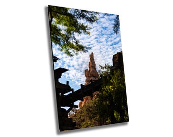 May include: A view of a tall, red rock formation with a wooden structure in the foreground. The sky is blue with white clouds.