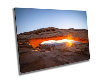 May include: A natural sandstone archway in a desert landscape with the sun rising behind it, creating a bright orange glow.