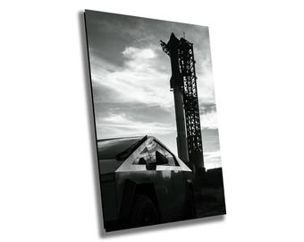 May include: A black and white photo of a futuristic-looking truck parked in front of a tall rocket launch tower. The truck is a pickup truck with a large, flat bed and a distinctive angular design. The rocket tower is a tall, metal structure with a large platform at the top. The photo is taken from a low angle, giving the viewer a sense of the truck's size and the tower's height.
