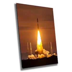 Puede incluir: Un lanzamiento de cohete al atardecer, con un cielo naranja brillante y una columna de humo y fuego que se arrastra detrás del cohete.