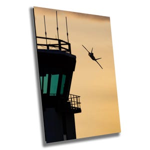 May include: Silhouette of a jet fighter plane flying past a control tower against a sunset sky.