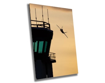 May include: Silhouette of a jet fighter plane flying past a control tower against a sunset sky.