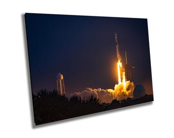 May include: A rocket launch at night with bright orange flames and smoke. The rocket is ascending into a dark blue sky with a few clouds. A water tower is visible in the distance.