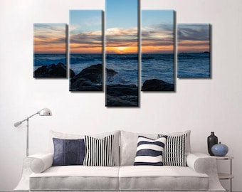 May include: A five-piece canvas print set depicting a seascape with a sunset over the ocean. The image features a blue sky with orange and pink clouds, a dark blue ocean with waves crashing on rocks, and a distant shoreline.