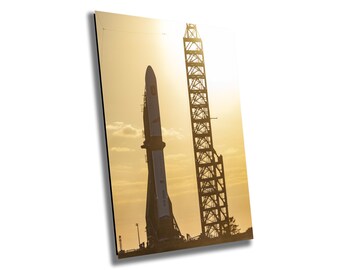 May include: A large rocket stands on a launchpad against a bright orange sunset. The rocket is silhouetted against the sky, with the sun shining through the structure of the launchpad.