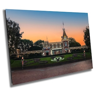 May include: A view of the entrance to Disneyland park at sunset. The iconic train station is visible in the background, with the words "Disneyland" and "Main Street, U.S.A." displayed on the building. The Mickey Mouse flower bed is in the foreground.