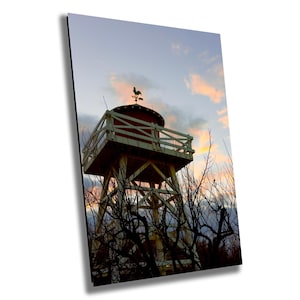 Puede incluir: Una torre de agua roja con una plataforma de madera blanca y una veleta de gallo en la parte superior. La torre se recorta contra un cielo nublado con una puesta de sol rosa y naranja.