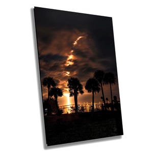 Op de afbeelding: Silhouetten van palmbomen tegen een donkere lucht met een heldere oranje en gele wolkenformatie. De stadslichten zijn in de verte zichtbaar.