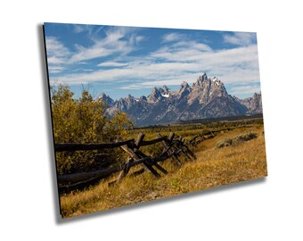 May include: A scenic landscape print featuring a wooden fence in the foreground, with a field of yellow grass and a mountain range in the background. The sky is blue with wispy clouds. The mountains have snow-capped peaks.